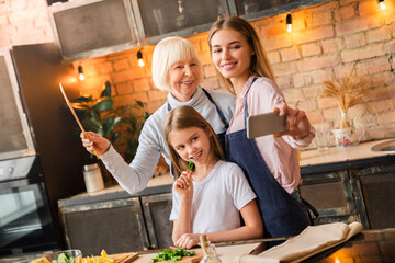 Happy young mother taking selfie with her daughter and grandmother in kitchen. Grandmother and...