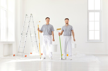 Portrait of smiling happy team of house painters looking at camera holding paint rollers and going to paint the wall of new apartment. Building contractors doing repair renovation preparing to move.