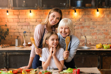 Horizontal front view shot of happy family that lookin in camera while cooking healthy food in kitchen. Grandmother hug her granddaughter with mother behind back. Happy family concept
