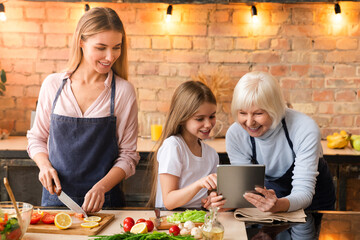 Cute little girl showing some to her mother and grandmother in the tablet while preparing fresh...