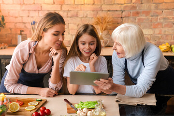 Front view of happy smiling family watching some information in the tablet while cooking food in kitchen. Granny mother and little daughter searching healthy meal recipe to internet