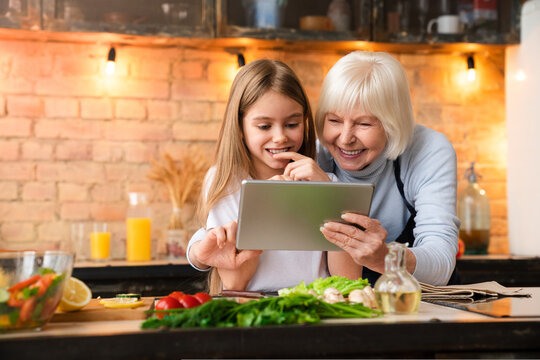 Grandmother with her little granddaughter searching in the tablet information while prepare salad in kitchen. Smiling senior woman and pretty girl using pad for cooking healthy food - Powered by Adobe