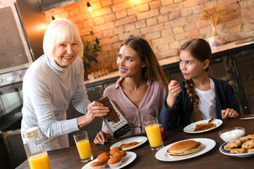 Side view of cheerful mature woman rubbing chocolate in the plate with pancakes for her daughter....