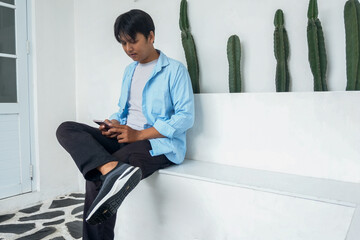 Confident Young Asian Male Using Mobile Phone While Sitting On Bench