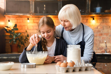 Cute little girl and her grandmother mixing batter in the glass bowl. Granny showing her teen...