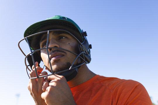 Indian man tightening green cricket helmet strap in orange athletic shirt on sports field