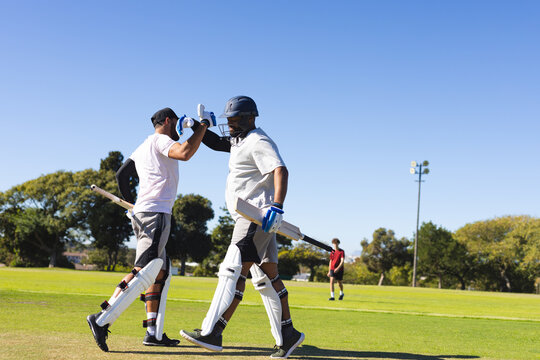 Diverse male cricket teammates walking on field high-fiving, holding bats in batting pads - Powered by Adobe