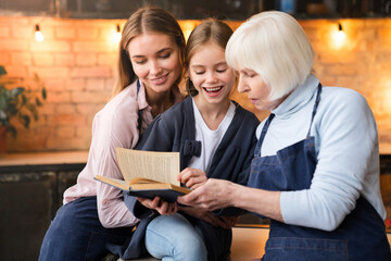 Happy three generations of women reading together recipe book in kitchen while cooking at home. Grandmother show her beautiful daughter and little grandchild secret family recipe