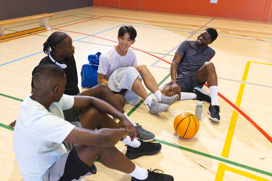 Diverse male teammates resting on basketball court with bag, water bottle, towel, headphones - Powered by Adobe