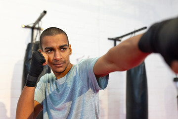Man training in boxing gym by punching bags in blue shirt and hand wraps punching forward