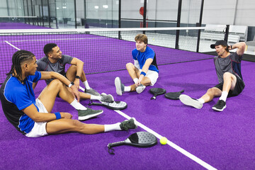 Diverse male friends sitting on purple padel court stretching with rackets, ball, watches