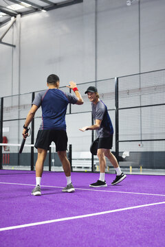 Two male teammates celebrating winning point on purple padel court by net with rackets, fence
