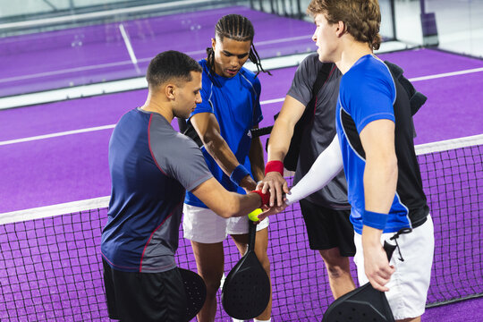 Diverse male teammates joining hands at net on purple padel court with rackets and padel ball