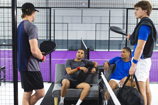 Diverse male friends resting on sofa chatting on padel court with racquets and bag with towel - Powered by Adobe