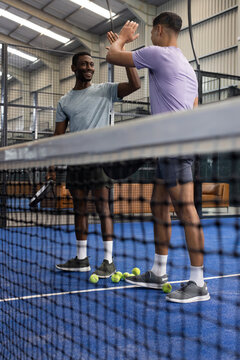 Diverse male teammates celebrating on blue padel court in sports facility with rackets, balls
