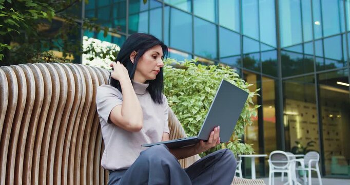 Concentrated young woman freelancer sitting on bench typing on laptop of background modern building. Serious pensive girl doing remote job. Outdoors