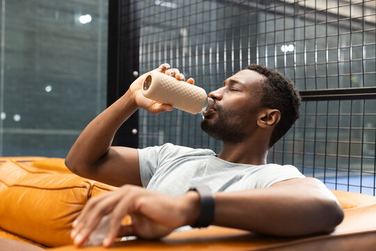 African american man sitting on orange bench in club lounge with reusable water bottle, watch - Powered by Adobe