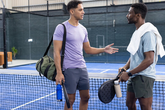 Diverse male friends in sportswear chatting at paddle tennis court net holding rackets, bottles - Powered by Adobe