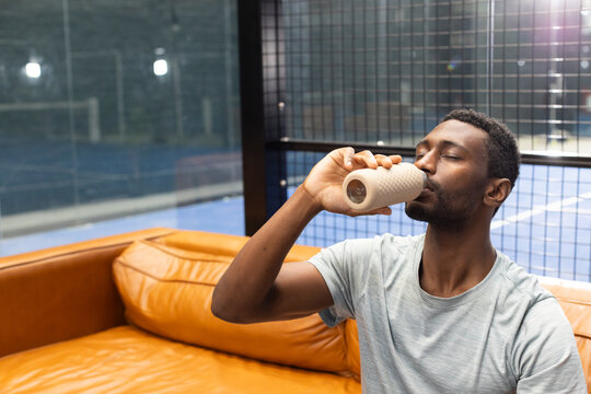African american man sitting on lounge couch in athletic tee drinking from water, copy space