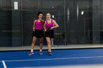 Diverse female teammates strategizing on padel court while adjusting racket grips next to wall
