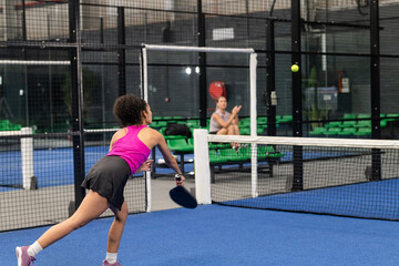 Diverse female training teammates lunging and hitting yellow padel ball with paddle on blue court