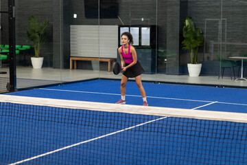 Female padel player standing on court with racket next to net, leaning forward in sports facility