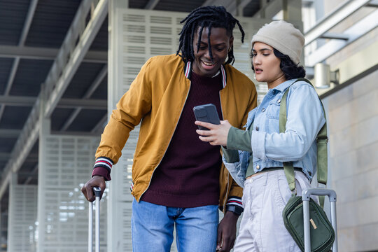 Diverse travellers pulling suitcases carrying crossbody bag, checking smartphone at station