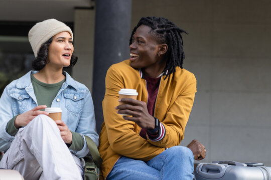 Diverse friends chatting and holding coffee at station bench near suitcase, messenger bag