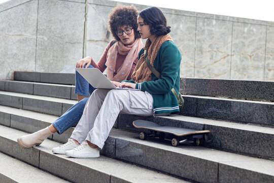 Diverse female friends sitting on steps in plaza using laptop near skateboard, shoulder backpack