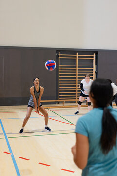 Diverse female volleyball teammates bumping tricolor volleyball on wood court wearing knee pads