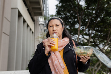 Woman standing outdoors at urban plaza holding orange juice cup, salad bowl, in polka dot scarf