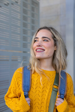 Woman standing on city sidewalk beside modern metal slat facade with denim backpack straps