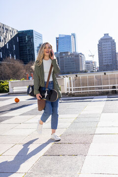 Female skateboarder walking on plaza, smartphone, passing bench rail, avoiding basketball