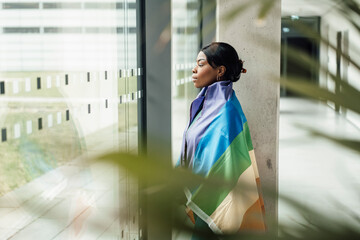 Young woman with rainbow flag looking out of window at office