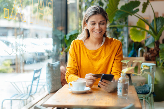 Happy woman holding credit card using smart phone in cafe