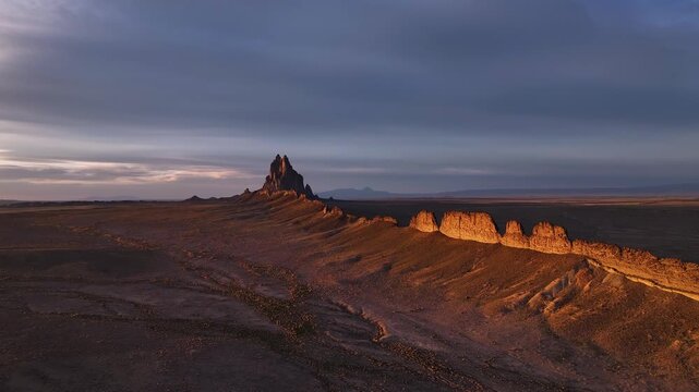 Aerial view of Shiprock's jagged silhouette piercing the horizon, casting long shadows across the arid landscape at dusk, Shiprock, New Mexico, United States.