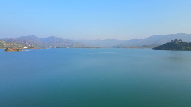 Aerial view of the still, turquoise waters of Khanpur Lake, contrasting with the rugged, brown mountains and green islands under a clear blue sky, Pakistan.