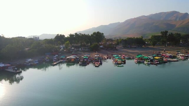 Aerial view of boats docked along the shore of the blue Khanpur Lake with mountains and trees in the background, Khanpur, Khyber Pakhtunkhwa, Pakistan.