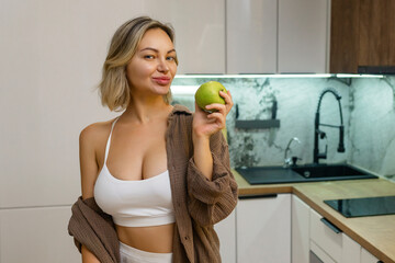 Attractive young woman in crop top playfully poses with a green apple in modern kitchen. Healthy...