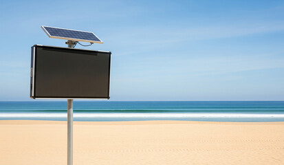 A black, solar-powered emergency call box or information kiosk stands on a beautiful, sunny beach, with the blue ocean and sky in the background.