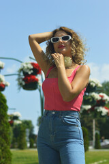 Caucasian slim blonde woman in pink top, blue jeans and white sunglasses walking along city street on summer sunny day.