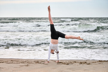 Young female in white top and black shorts performs cartwheel on sandy baltic sea beach. Overcast...