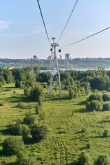 Obraz premium Cableway travels above lush green landscape toward distant city skyline. Clear blue sky and river create serene atmosphere. Captured from elevated perspective