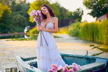 Beautiful woman in white dress on a boat with flowers on a lake in the evening light.