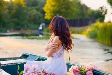 Beautiful woman in white dress on a boat with flowers on a lake in the evening light.
