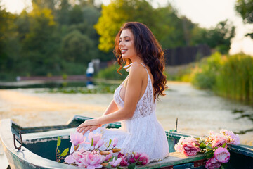Beautiful woman in white dress on a boat with flowers on a lake in the evening light.