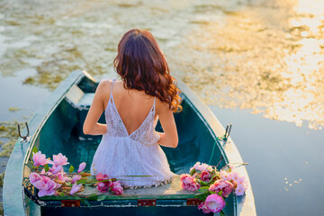 Beautiful woman in white dress on a boat with flowers on a lake in the evening light.