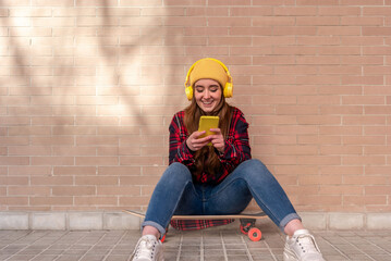 Happy student girl using smartphone and listening music with headphones while sitting on skateboard
