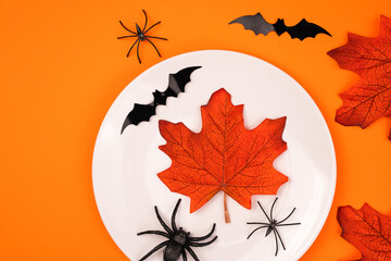 White plate with autumn maple leafs, spiders and black bats on bright orange background. Halloween party and table setting concept. Flatlay. Top view. Copy space.