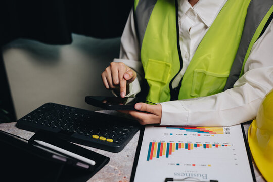 Business Professional Analyzing Data on Smartphone in Safety Vest and Hard Hat at Office Desk with Reports and Keyboard